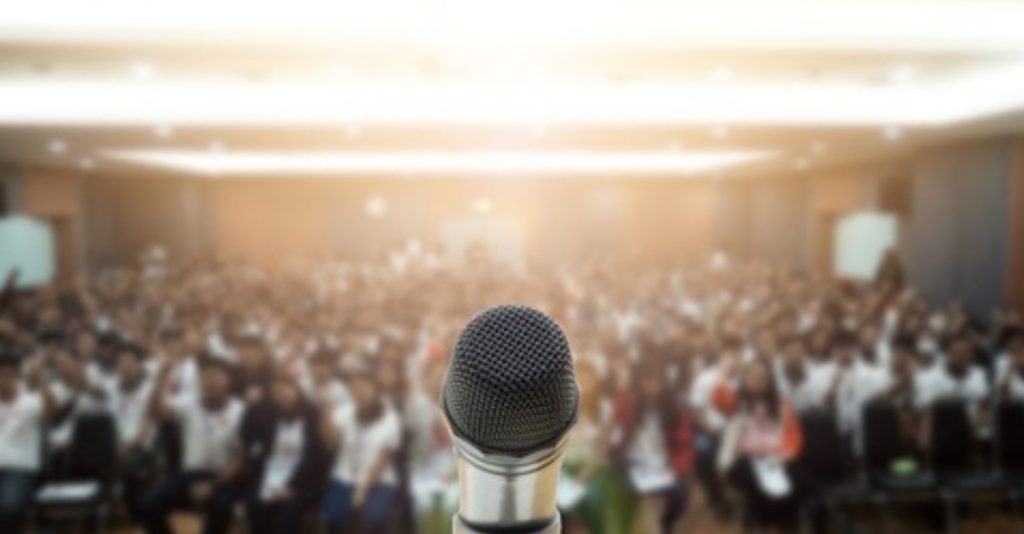 Microphone in focus against a blurred backdrop of an engaged audience at a Sava Politis keynote speech, symbolising empowerment and inspiration.