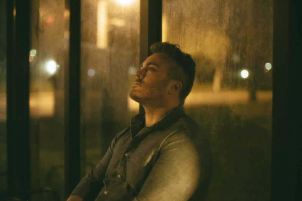 Man sitting alone in quiet reflection at night, gazing upward through a window, evoking a sense of emotional heaviness and inner searching.